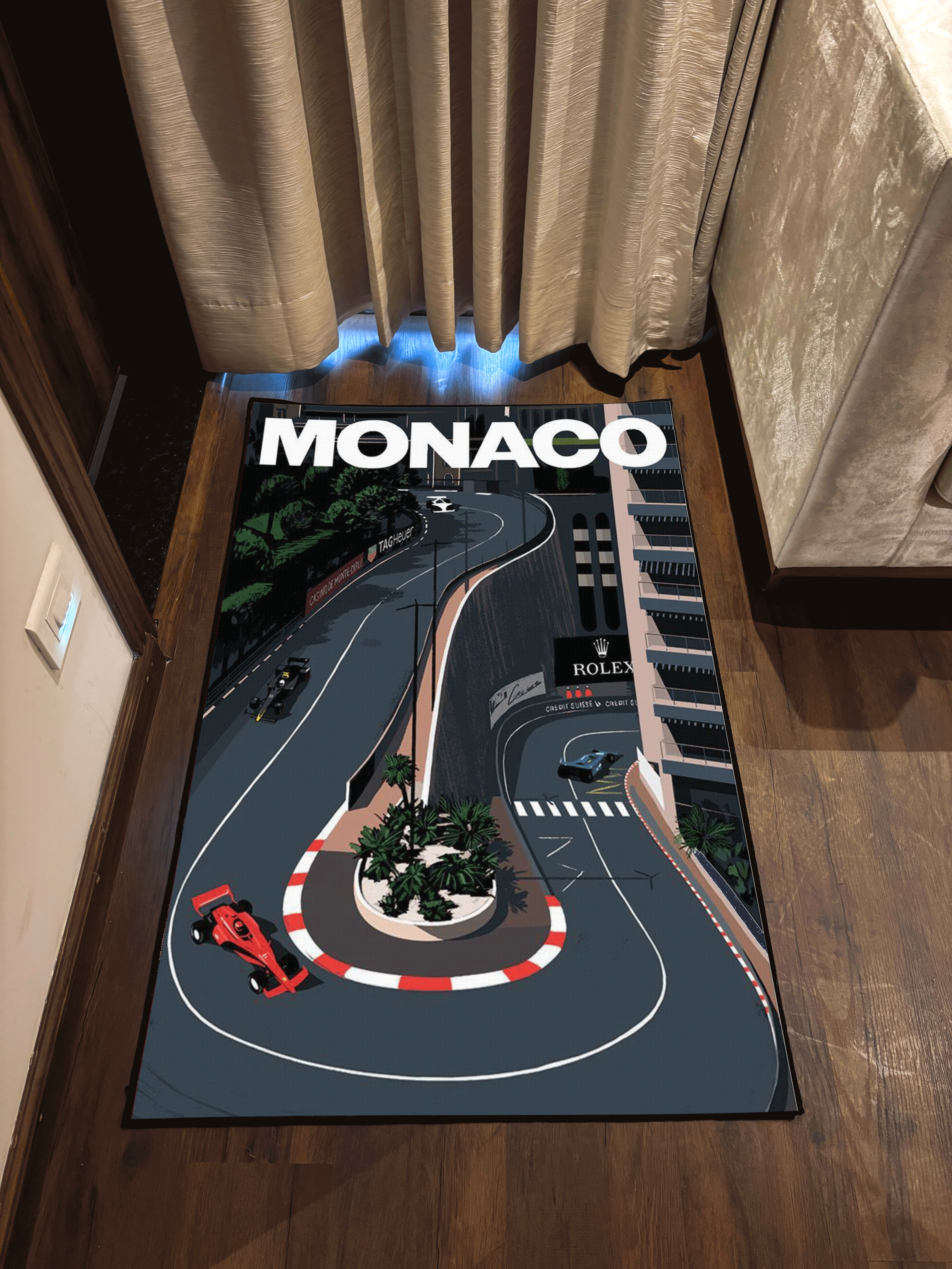 A washable rug with a Monaco F1 racing track print, placed on a wooden floor near a curtain, with a car and track illustration on the cover.